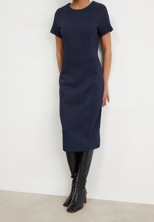 Frau trägt ein tailliertes, kurzärmeliges marineblaues Midikleid kombiniert mit schwarzen kniehohen Stiefeln und steht vor einer weißen Wand.