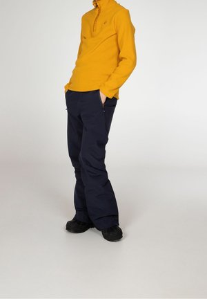 Pullover de forro polar amarillo con media cremallera combinado con pantalones azul marino. El pullover incluye un cuello y los pantalones tienen bolsillos con cremallera. Calzado negro.