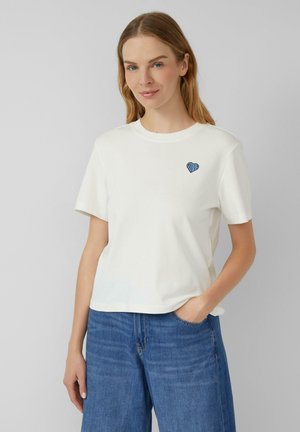 T-shirt en coton blanc à manches courtes, col rond, avec un motif de cœur rayé bleu sur le côté gauche de la poitrine. Texture lisse.
