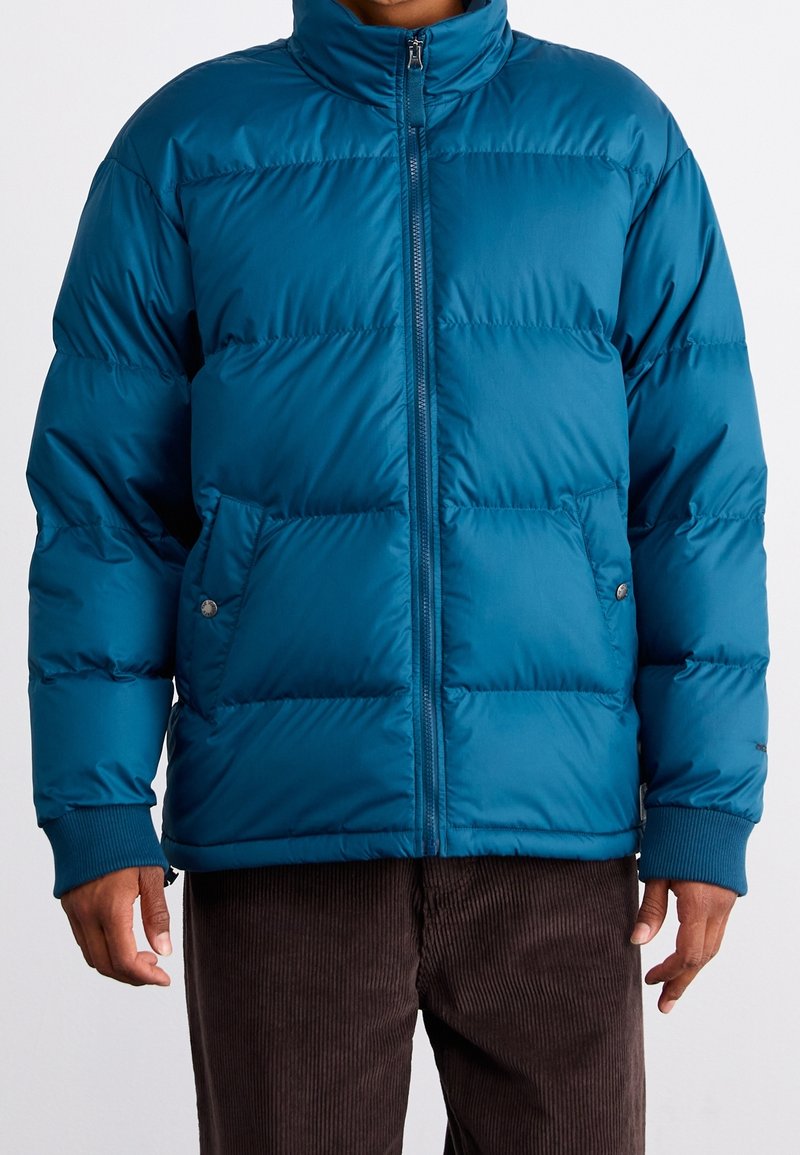 Turkos vadderad jacka med hög krage, dragkedja och två framfickor. Har vadderade partier och ribbade muddar. Mjuk, syntetisk yta.