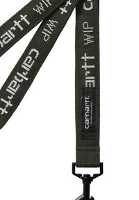 Correa de tela verde tejida con el texto "carhartt WIP" en blanco, con una etiqueta rectangular negra y un clip en un extremo.