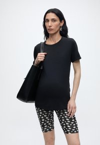 Femme aux longs cheveux foncés portant un t-shirt noir, un short noir à motifs floraux, des boucles d’oreilles en argent et portant un grand sac à main noir sur l’épaule.