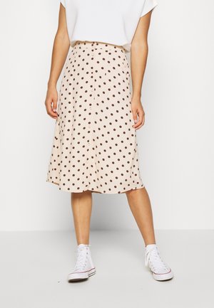 Femme portant une jupe midi beige à pois marron, un haut blanc à manches courtes et des baskets montantes blanches, debout contre un mur uni.