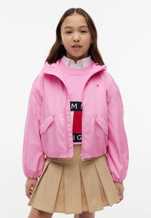 Pige iført en pink hættejakke over en pink strikket trøje med logo, hvid skjorte med krave og beige plisseret nederdel, stående mod hvid baggrund.