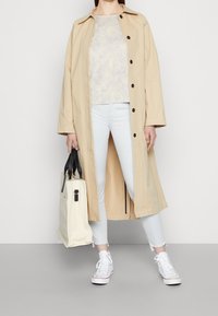 Béžový lehký trenčkot s černými knoflíky, vzorovaným topem, svetle modrými džínami a bílými teniskami, doplněný krémovou taškou přes rameno s černými uchy.