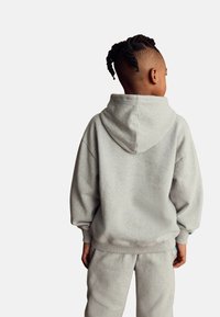 Sudadera de felpa gris con un bolsillo tipo canguro y capucha ajustable, combinada con pantalones de chándal gris a juego. Presenta puños y dobladillo acanalados.