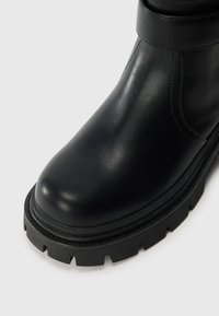 Close-up of black leather boot with rounded toe and thick, rugged rubber sole on white background.
