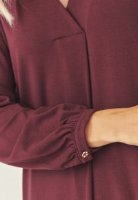 Femeie îmbrăcată cu o bluză maronie cu mânecă lungă, decolteu în V și manșete cu nasturi, arătând o parte din brațul și trunchiul său.