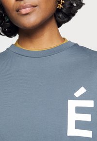 Sininen puuvillaneule, jossa on edessä valkoinen "É"-logo. Alapuolella näkyy kerroksesta peräisin oleva keltainen kaulus. Pehmeä tekstuuri.