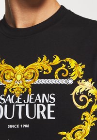 Sort t-shirt med udsmykkede gyldne barok-illustrationer og hvid tekst, der står "VERSACE JEANS COUTURE" med "SINCE 1988" nedenunder.