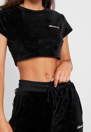 Vrouw draagt een zwart cropped t-shirt en zwarte broek met trekkoord en ellesse-logo op de borst en broekspijp.