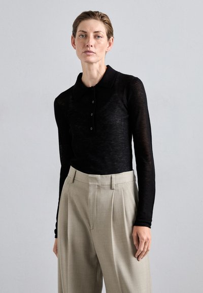 Polo shirt noir à manches longues avec un col et une fermeture à boutons, associé à un pantalon beige clair plissé. Texture de tissu lisse.