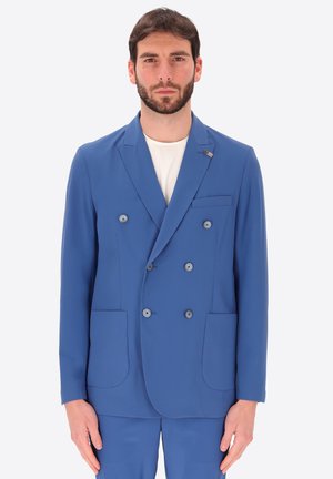 Uomo con la barba che indossa una giacca blu doppiopetto sopra una camicia bianca, in piedi di fronte a uno sfondo chiaro e semplice.