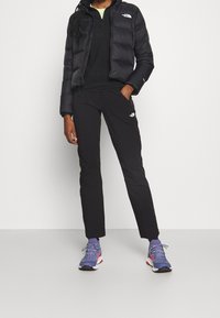 Veste en duvet noire North Face, haut noir, pantalon noir et chaussures de sport violettes. La veste présente un design matelassé et une fermeture éclair ; les pantalons ont une coupe slim.