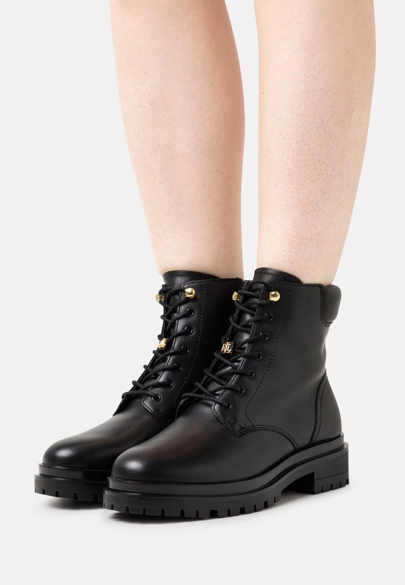 Black leather ankle boots with gold hardware, round toe, lace-up design, and textured rubber sole, showcasing a sleek, modern shape.