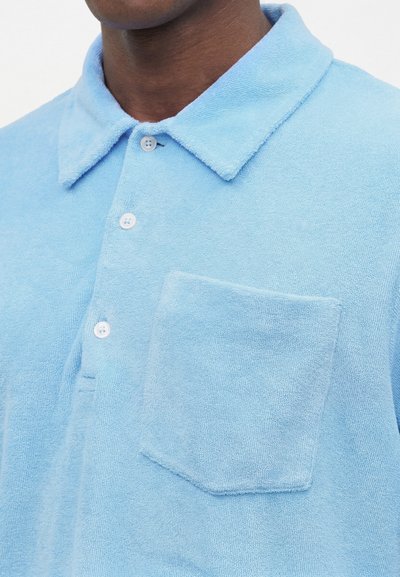 Κοντινό πλάνο ενός ατόμου που φορά ένα φωτεινό γαλάζιο πόλο από πετσέτα με γιακά, τρία κουμπιά και μια τσέπη στο στήθος αριστερά.