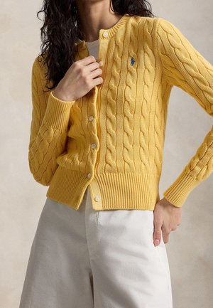 Femme portant un cardigan en maille torsadée jaune avec des boutons blancs et un pantalon blanc, main ajustant le cardigan près du col.