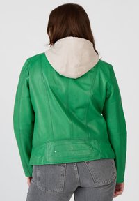 Grön läderjacka med en beige huva. Har strukturerade sömmar och en slät yta. Följt av grå högmidjade jeans.