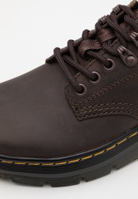 Brown leather shoe with a rounded toe, textured surface, round laces, reinforced eyelets, and contrasting yellow stitching on a black sole.