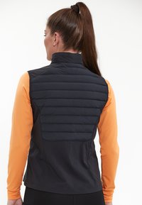 Schwarze isolierte Weste mit hohem Kragen, ausgestattet mit einem gesteppten Rücken und einer glatten Vorderseite. Das Modell trägt darunter ein orangefarbenes Langarmshirt.
