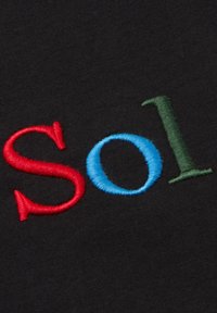Schwarzer Stoff mit gesticktem Text "Sol" in roten, blauen und grünen Farben. Die Nähte sind sichtbar und verleihen eine strukturierte Optik.