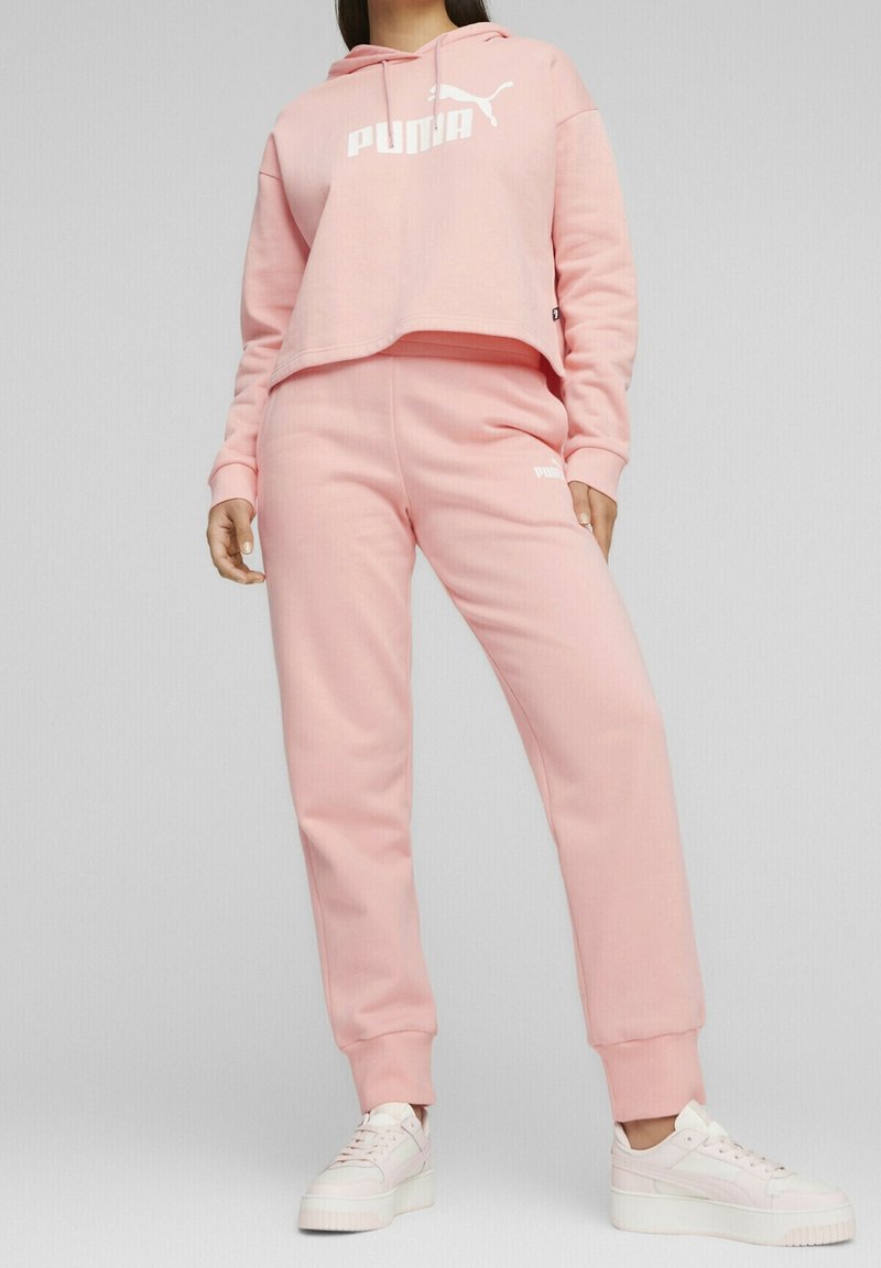 Roze cropped hoodie met witte "PUMA" logo, gecombineerd met bijpassende roze joggers. De stof lijkt zacht. Witte sneakers maken de outfit compleet.