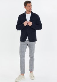 Blazer azul marino con dos bolsillos frontales, dos botones y solapas con muesca. Combinado con pantalones de cuadros gris claro y zapatillas beigas.