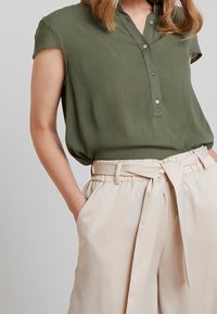 Blusa de manga corta en verde oliva con una placket de botones, combinada con pantalones de tiro alto en beige que cuentan con una cintura ceñida y bolsillos laterales.