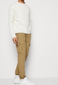 Pull en maille blanc, pantalon cargo beige avec poches latérales et baskets blanches. Look décontracté avec des détails côtelés et des poignets élastiques.