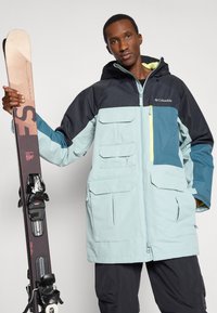 Kurtka narciarska w jasnym niebieskim i granatowym kolorze, z wieloma kieszeniami, zamkiem błyskawicznym i kapturem. Model trzyma narty z wiązaniami.
