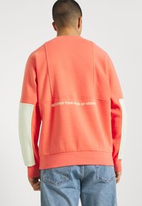 Sweatshirt corail avec des manches contrastantes vert clair. Comprend un slogan "DÉPASSEZ VOTRE PEUR DES HAUTEURS" centré au dos. Texture douce.