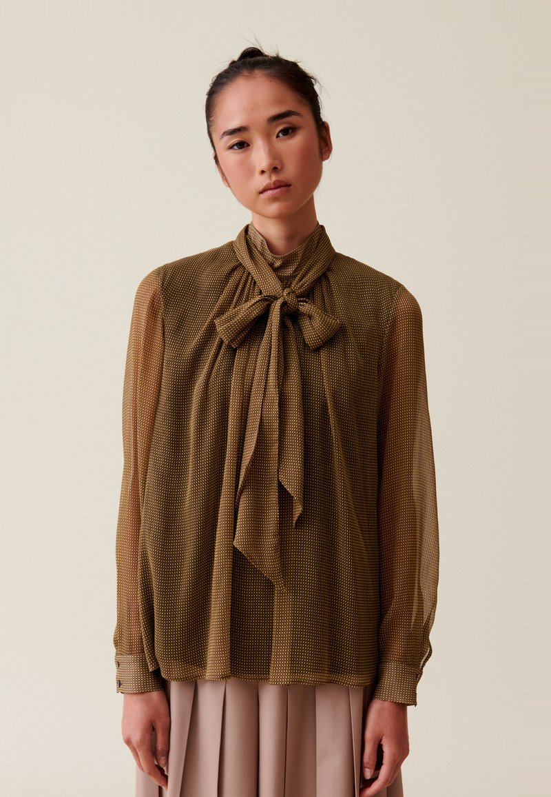 Mujer vistiendo una blusa oliva clara y transparente con un gran lazo en el cuello y una falda plisada beige, de pie contra un fondo liso.