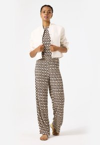 Crèmekleurige cropped jas van textuurstof, gedragen over een gestreepte jumpsuit met diamantvormen in beige en bruin. Tan sandalen maken de look compleet.