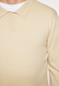 Beige katoenen polo shirt met lange mouwen en een kraag, voorzien van een knoopsluiting en drie knopen. Soepele textuur met ribbeldetail.