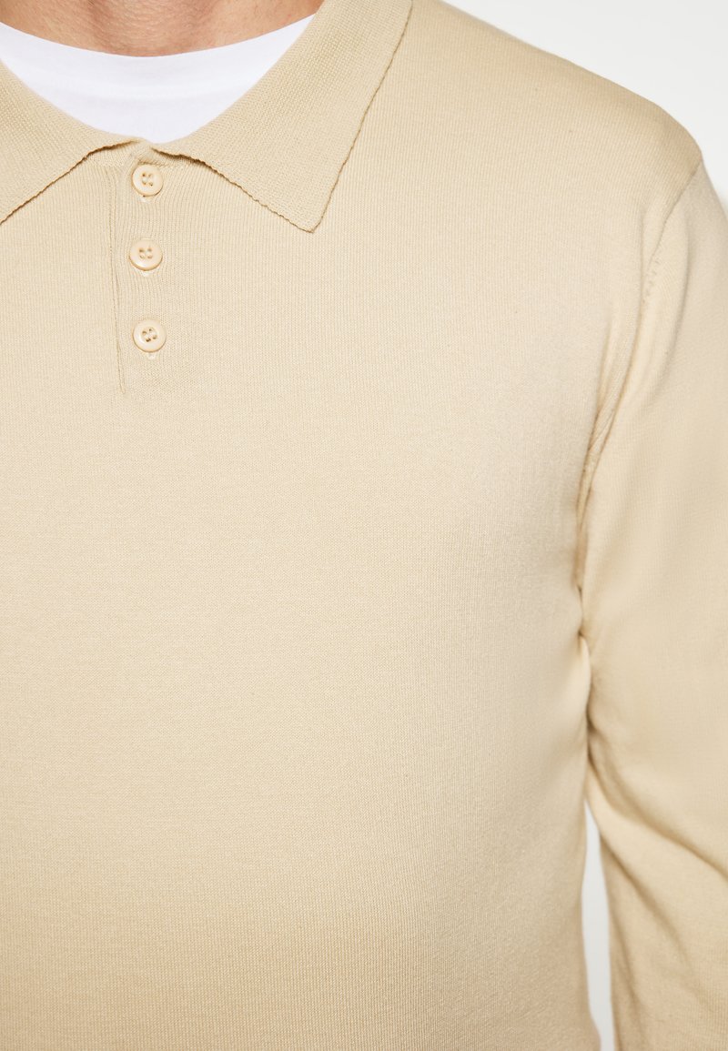 Beige katoenen polo shirt met lange mouwen en een kraag, voorzien van een knoopsluiting en drie knopen. Soepele textuur met ribbeldetail.