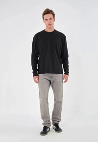 Zwarte langemouwen shirt gecombineerd met lichtgrijze jeans en zwarte sneakers, staand tegen een effen witte achtergrond.