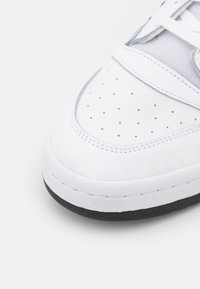 Hvid sneakers med en glat læderoverdel, perforeret tåboks og teksturerede grå detaljer. Den har en sort gummisål og hvide snørebånd.
