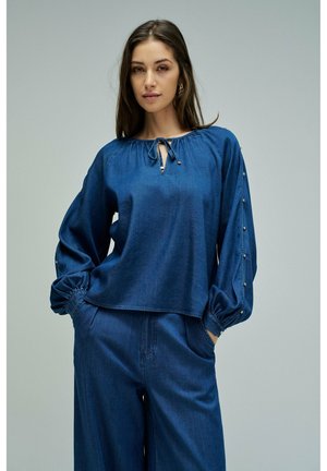 Femme portant un chemisier en denim bleu marine ample avec des détails de boutons sur les manches et un pantalon en denim assorti, debout les mains dans les poches.