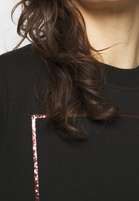 Černá mikina s lesklým, růžově zlatým obdélníkovým akcentem. Měkká textura a žebrovaný kulatý výstřih. Vlasy částečně viditelné na jedné straně.