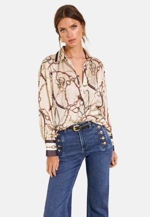 Vrouw draagt een crèmekleurige satijnen blouse met kettingprint en omgeslagen mouwen, gecombineerd met een high-waisted blauwe spijkerbroek met gouden knopen.