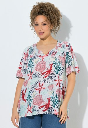 Mujer con cabello rizado lleva una camiseta colorida de manga corta con patrones de estrellas de mar rojas, conchas marinas y algas, y vaqueros azules sobre un fondo blanco.