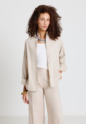 Femme aux cheveux bouclés portant une chemise beige boutonnée, un pantalon large assorti, un haut blanc, des colliers superposés et un bracelet épais, debout en position neutre.