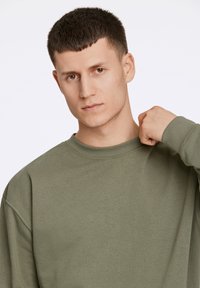 Sudadera holgada de mangas largas en color verde oliva con cuello acanalado. Tela suave con una textura sutil, que presenta costuras de hombro caídas.