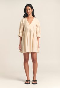Beige katoenen minidress met een V-hals, knoopdetails en pofmouwen. Beschikt over een gathered taille en een uitstaande zoom; gedragen met zwarte sandalen.