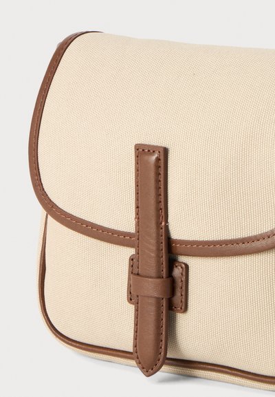 Sac bandoulière en tissu beige avec garniture en cuir marron et fermeture à sangle frontale sur fond uni.