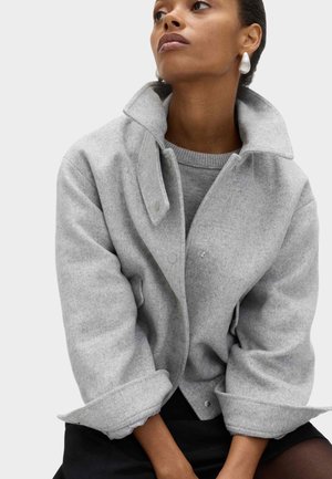 Chaqueta gris de mezcla de lana con cuello alto, corte holgado y cierre de botones. Lleva encima un suéter gris ligero. Diseño minimalista.