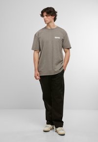 Tricou din bumbac gri, cu mâneci scurte, având un imprimeu pe partea din față. Asortat cu pantaloni negri și adidași albi. Fundal simplu.
