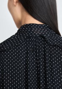 Schwarze Bluse aus strukturiertem Stoff mit weißen Polka-Dots, einem gewellten Kragen und einem kleinen Knopfverschluss auf der Rückseite.