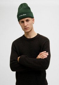 Zielona dzianinowa czapka beanie o wzorze prążkowanym z białym logo "JJ studios". Ciemny sweter teksturowany z okrągłym dekoltem i luźnym krojem.