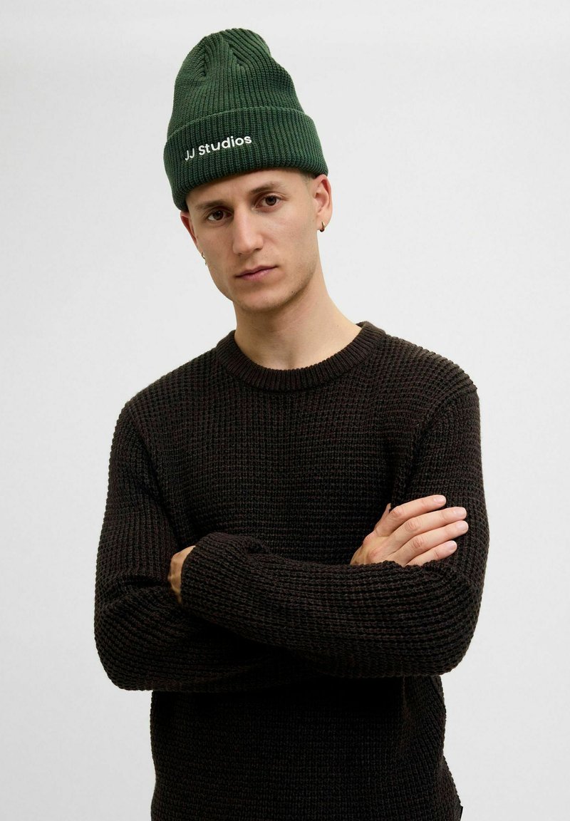 Zielona dzianinowa czapka beanie o wzorze prążkowanym z białym logo "JJ studios". Ciemny sweter teksturowany z okrągłym dekoltem i luźnym krojem.